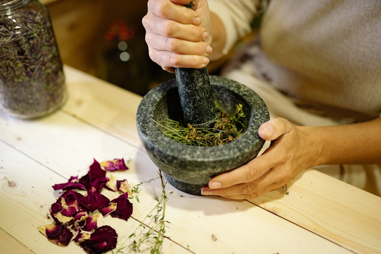 about-img-01 A person grinding herbal ingredients in a stone mortar and pestle with dried petals nearby.