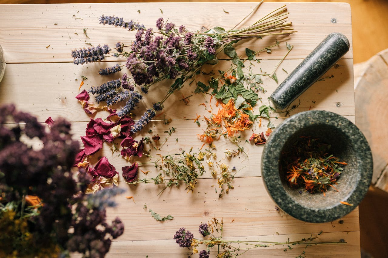 gallery-03 Flat lay of dried herbs and flowers with a stone mortar and pestle on a rustic wooden table.